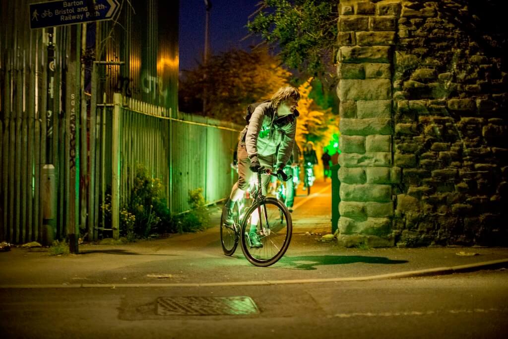 Cyclists emerge from an alley riding bikes adorned with bright green lights.