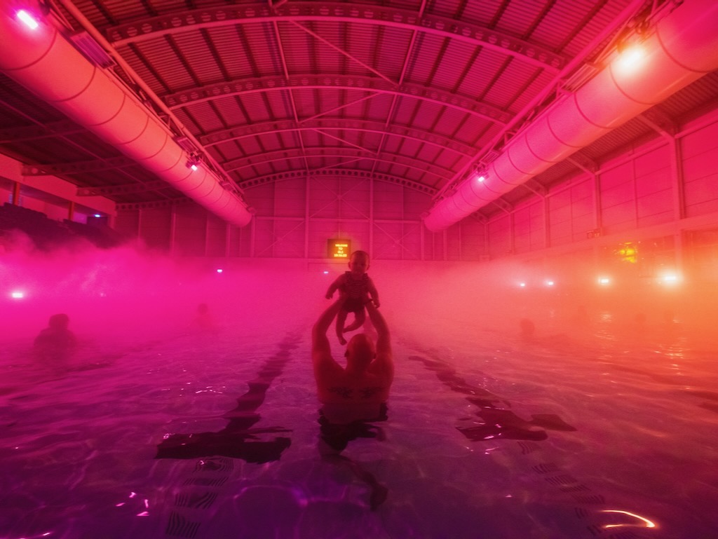 A father raises his baby into the air inside a pink swimming pool surrounded by ethereal pink fog.