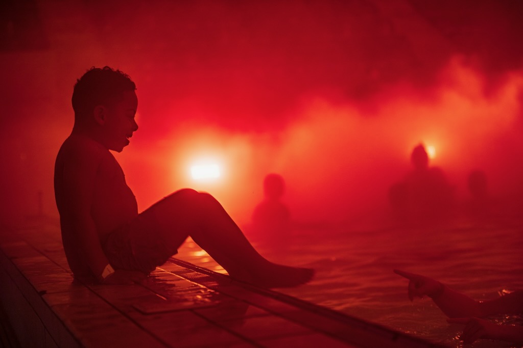 A child laughs with joy on the edge of a swimming pool enshrouded by bright red fog.