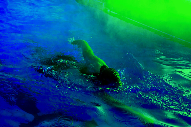 A swimmer creates colourful ripples as the pool reflects bright blue and green fog.