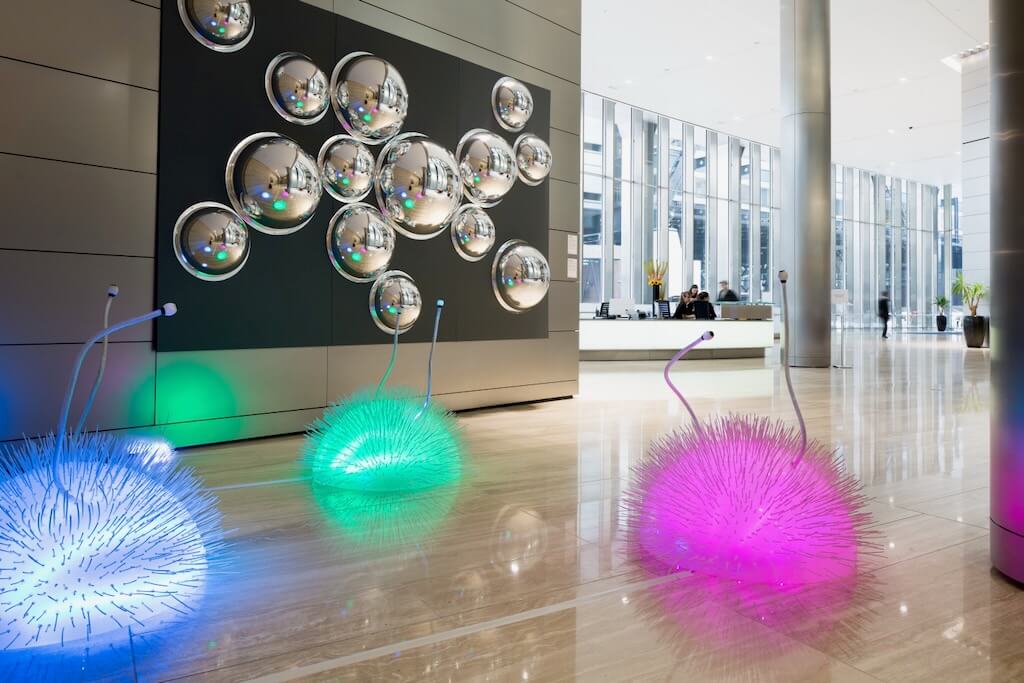 3 spiky creatures glowing pink, green and blue sit underneath reflective silver orbs mounted on a wall.
