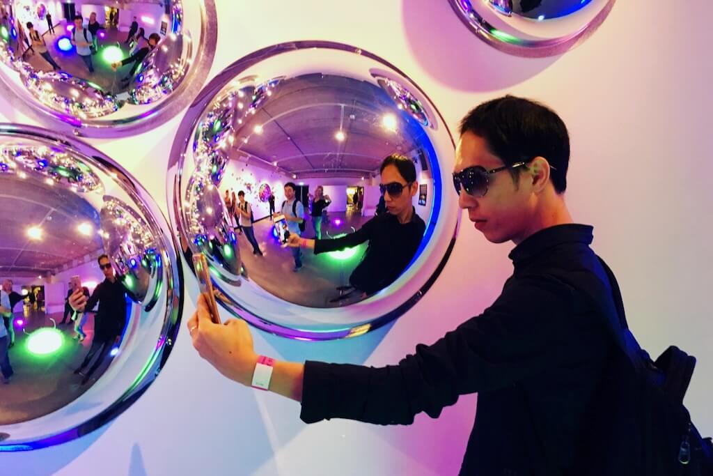 A man poses for a selfie with his warped reflection in a silver orb.