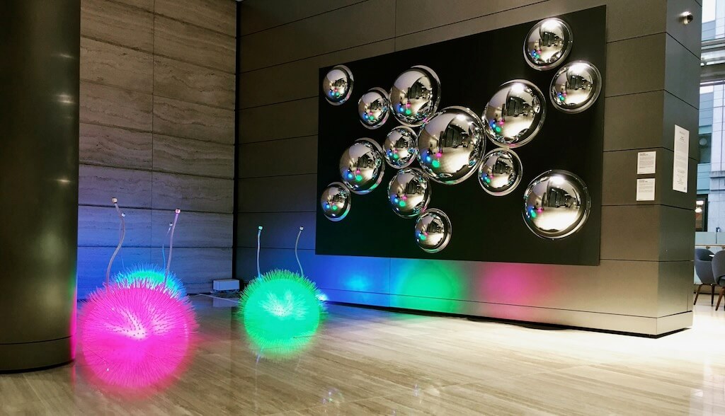 3 spiky creatures glow pink, green and blue underneath reflective silver orbs mounted on a wall.