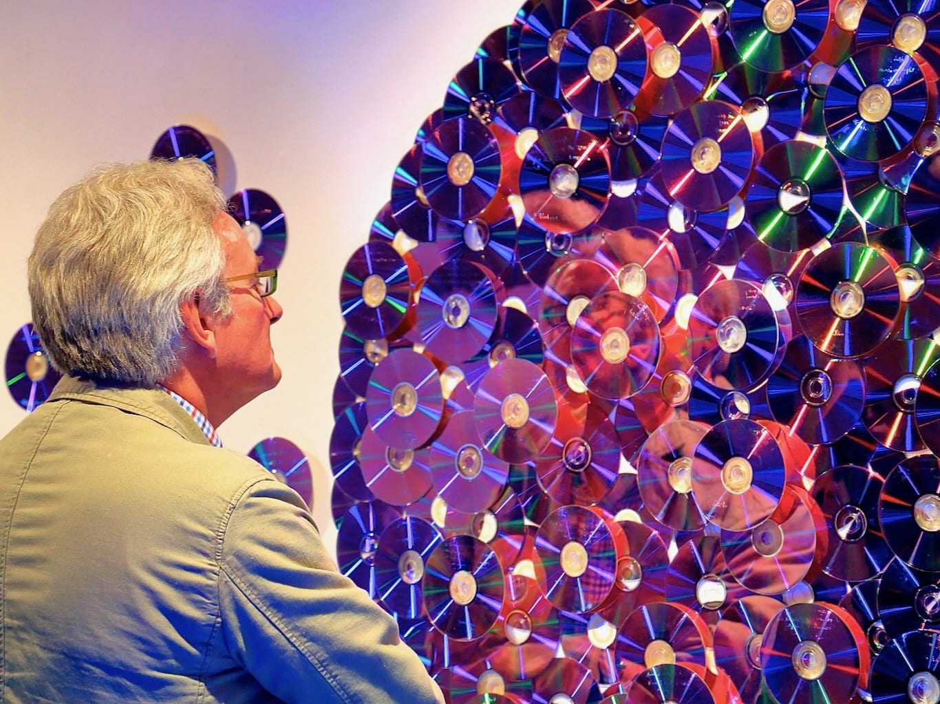 An older man studies his abstract reflection created by layers of reflective disks mounted in a circular formation.