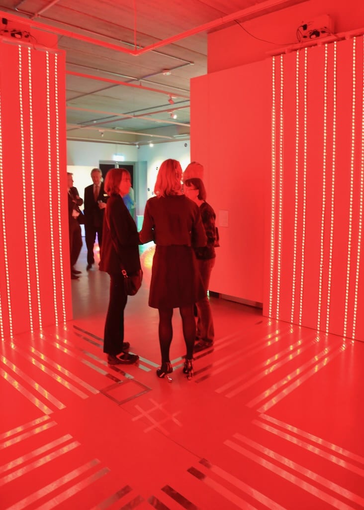 Ribbons on red light on the walls are reflected by opalescent strips running perpendicular across the floor.