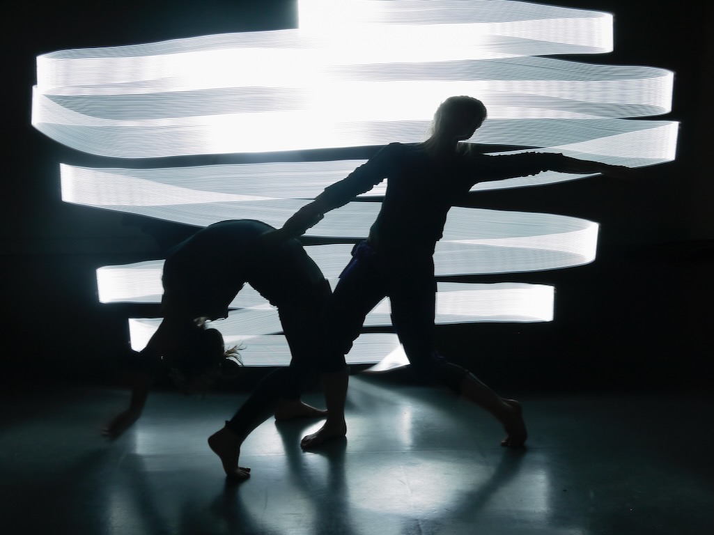 The silhouettes of two dancers are illuminated by ribbons of white light.