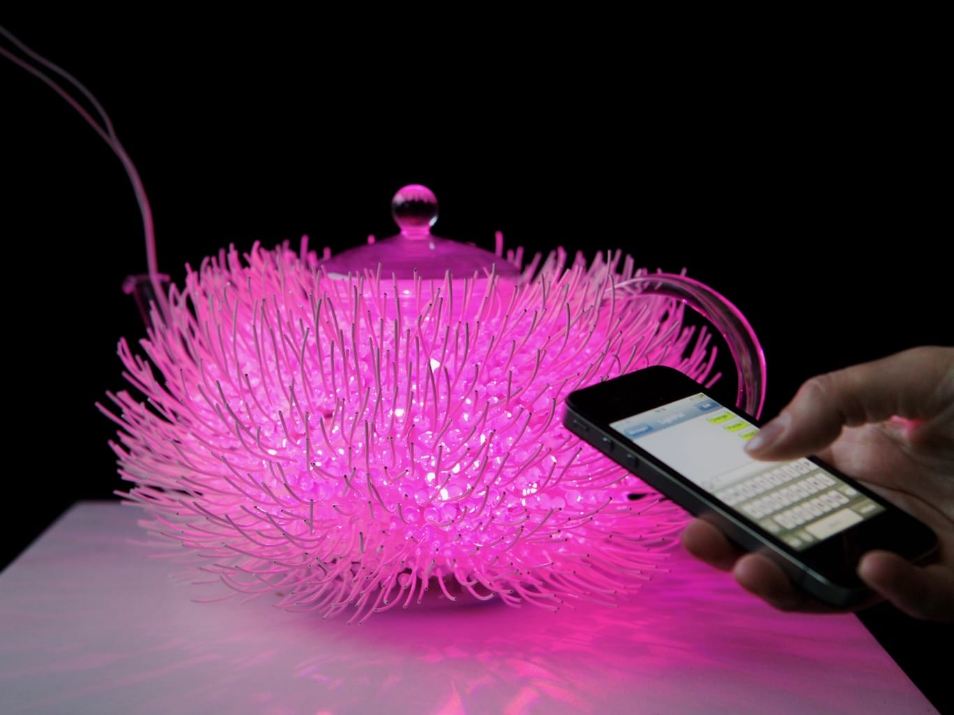 A person uses their phone to activate different colours shining through the teapot.