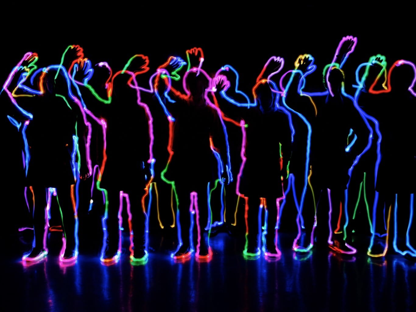 A group of silhouettes outlined in a rainbow of lights wave hello.