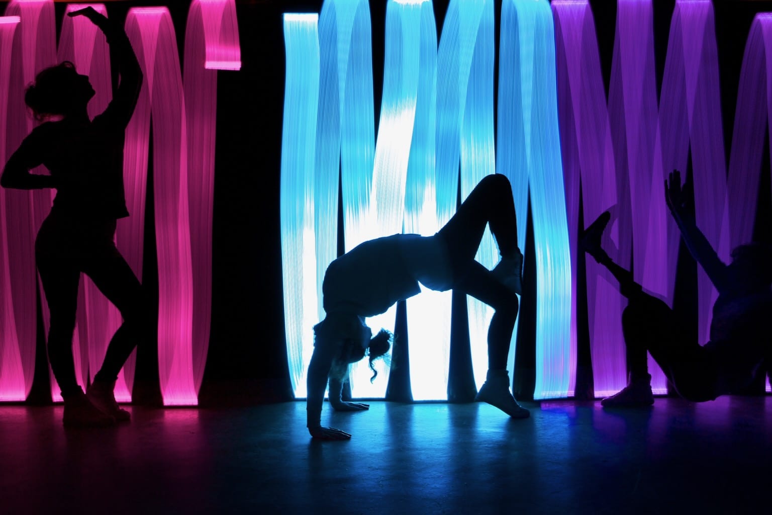 3 young dancers are illuminated by ribbons of colourful light, each striking a different pose.