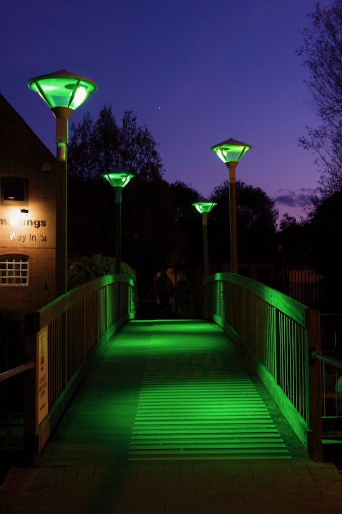 Motion sensing lampposts illuminate a wooden bridge with vibrant green hues.