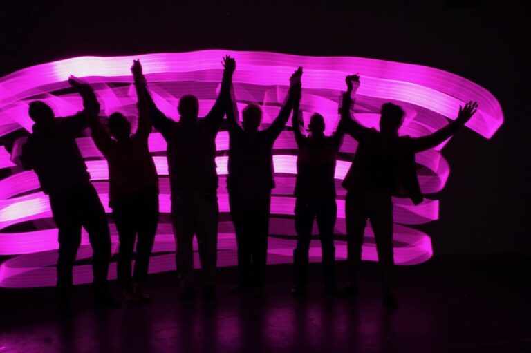 6 joyful silhouettes are illuminated by pink ribbons of light.